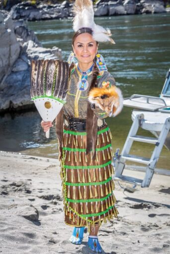 A woman in traditional indigenous attire holding a fan stands by a riverbank in Iconic Idaho with a small dog and a boat in the background.