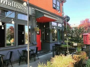 Exterior of "mulleady's" pub with vibrant orange walls, hanging flower pots, outdoor seating, and string lights, featured in Seattle magazine.