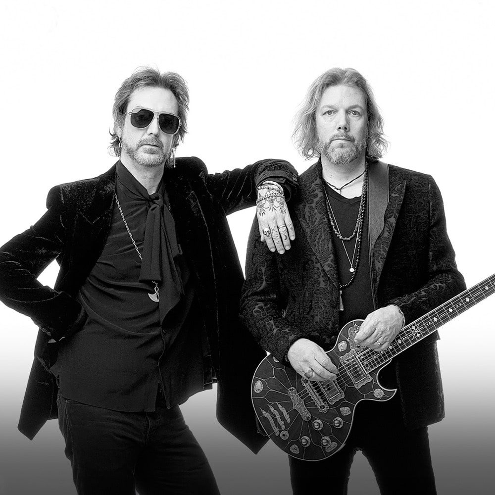 Two men in black attire and sunglasses posing with one holding a guitar, listed as a must-see for April 11-17.