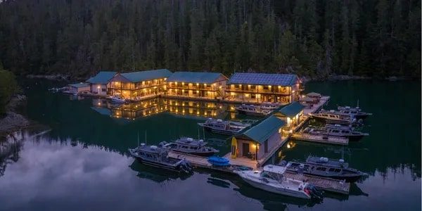 Illuminated culinary retreat lodge with adjacent docks and boats on a serene lake surrounded by forest at dusk.