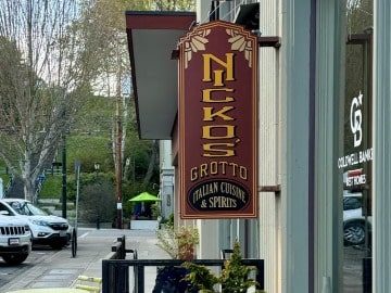 A vertical street sign for "Nick's Grotto Italian Cuisine & Culinary Retreat" mounted on a post, with a street and greenery in the background.