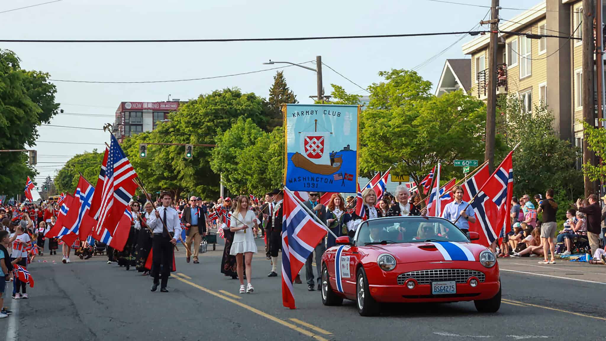 A parade celebrating Norwegian Constitution Day features people marching with Norwegian flags. A red convertible decorated with flags leads, followed by a banner for the Leif Erikson Lodge 2-001 and other participants on a street lined with spectators. Hip hurra echoes through the crowd, making it a festive Friday event.