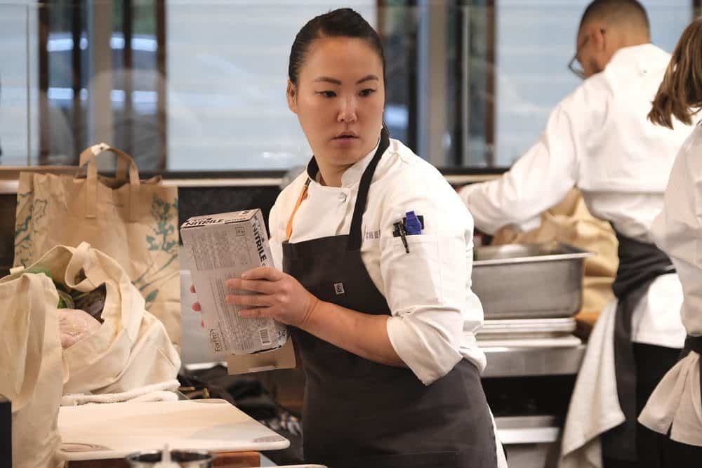 A focused female chef, Kaleena Bliss, wearing a white apron reads a "Cream of the Chop" carton while working in a busy "Top Chef" kitchen environment.