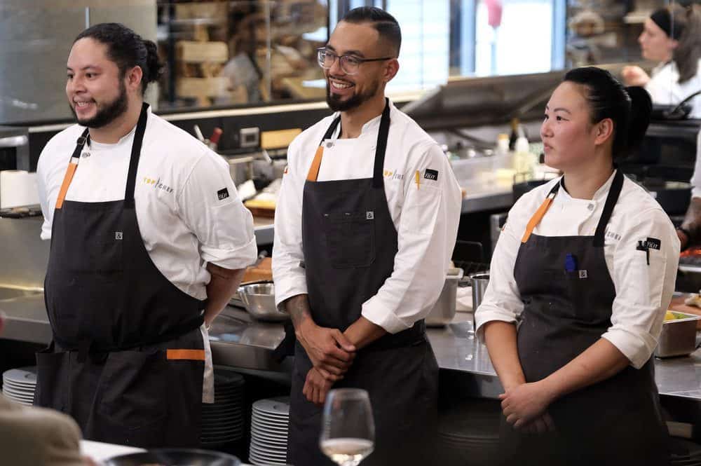 Three chefs in uniform, including Kaleena Bliss, are standing in a commercial kitchen environment.