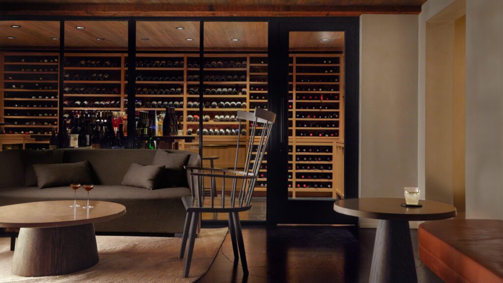 Interior of a sophisticated bar with dark wood furnishing, round farm tables, and a wall-mounted wine rack, dimly lit for ambiance.