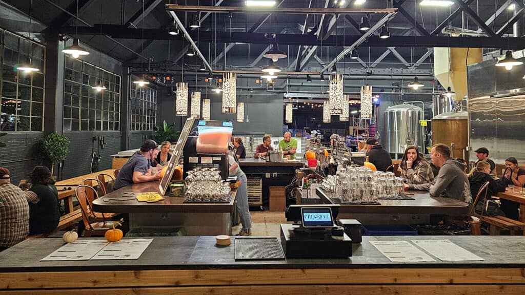 A spacious brewery taproom with happy patrons seated at tables and the bar, illuminated by pendant lights. Large brewing equipment is visible in the background, hinting at the hoppy creations made on-site.