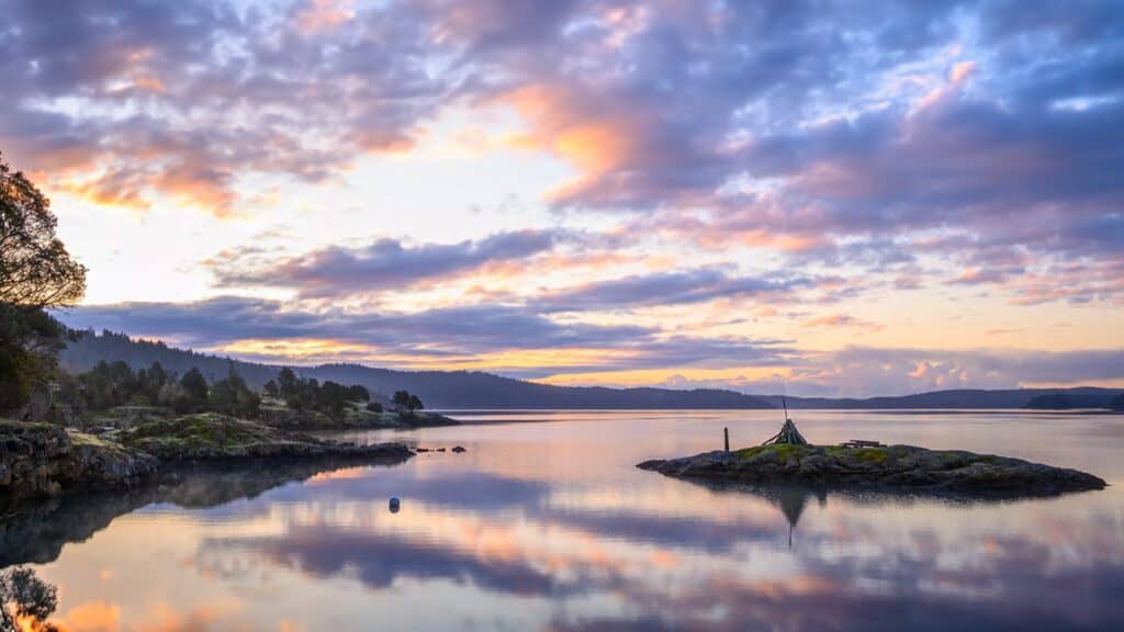 Tranquil landscape at sunrise featuring a reflective lake, colorful sky, and a small island connected by a narrow strip of land, ideal for island hopping.