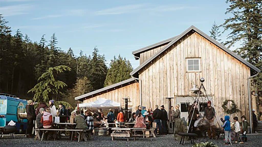 A rustic barn gathering with people socializing outdoors, surrounded by picnic tables and forest under a clear sky, perfect for island hopping enthusiasts looking to experience local tourism.