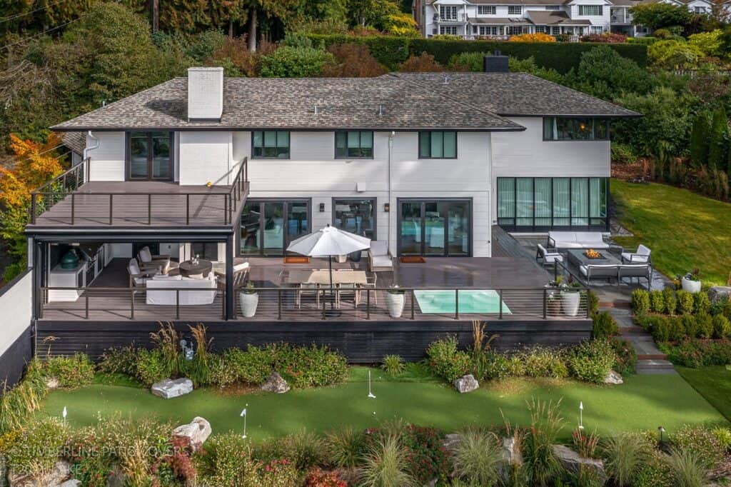 Aerial view of a two-story modern house with a large deck, outdoor seating, a small pool, and a putting green, surrounded by lush greenery and other houses in the background. Sponsored by People Come First (PCF), this home epitomizes luxury living close to nature.