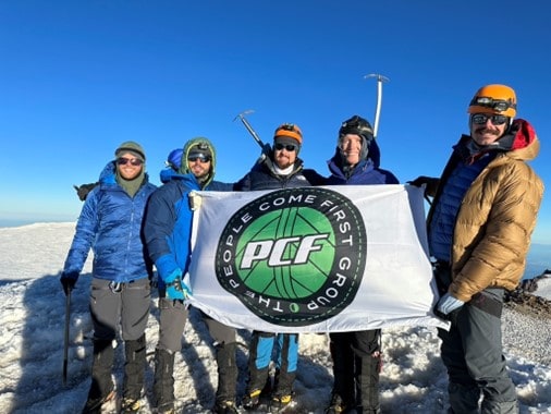 A group of five people in winter gear stand on a snowy mountain peak holding a banner that reads "PCF: People Come First Group.