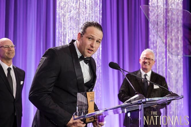 A man in a tuxedo stands at a podium holding an award, sponsored by PCF, speaking into a microphone. Two other men in suits smile in the background. The backdrop is purple with vertical light decorations.