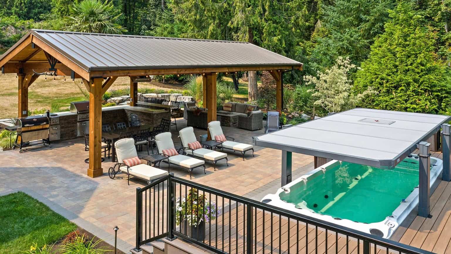 A backyard with a covered patio, sponsored by PCF, featuring a dining area and lounge chairs next to a swimming pool with a retractable cover, surrounded by trees and greenery.