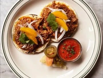 Three chicken tacos topped with onion and cilantro, featured in Seattle magazine, served with a side of salsa, pickled vegetables, and a slice of lime on a white plate.