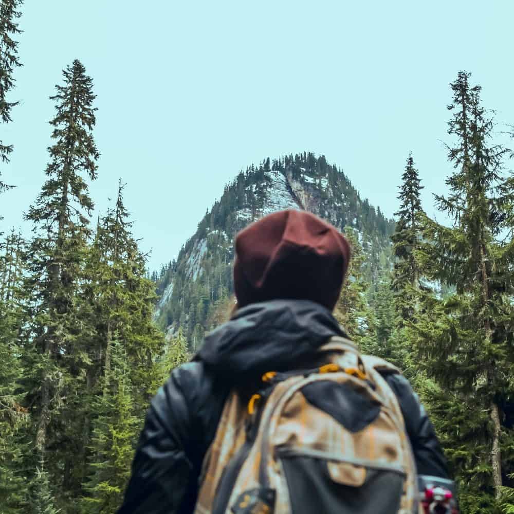 A person wearing a backpack facing a forested mountain under a cloudy sky. Must List, April 18-24.