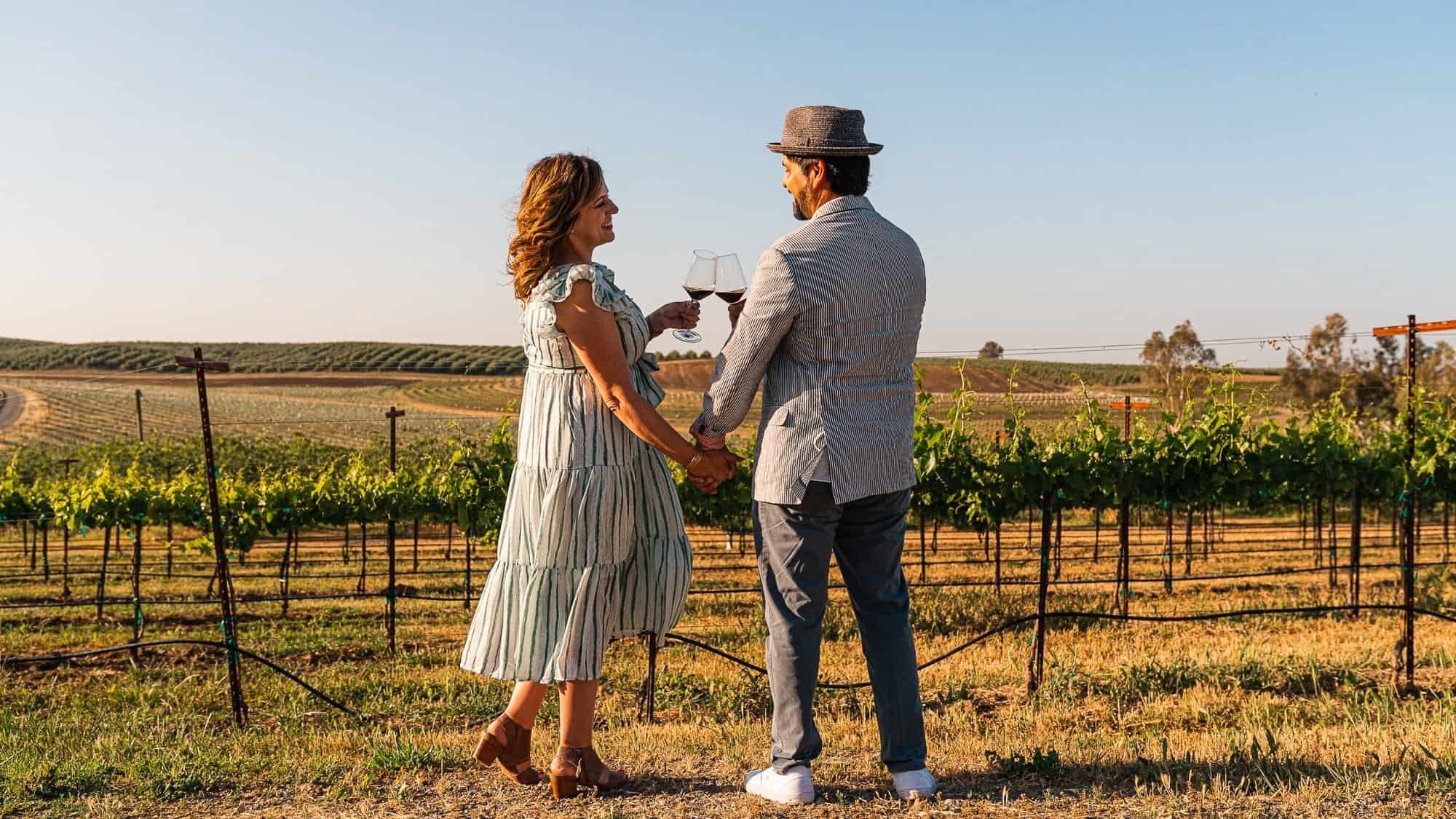 Couple holding hands and toasting with wine in a hidden gem vineyard at sunset.