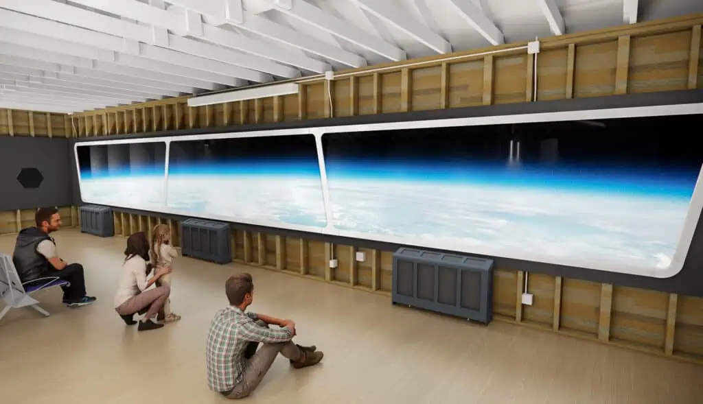 A group of people sits and kneels in a room with a large window showing a view of Earth from space. The interior, part of the Museum of Flight's new exhibit on Life in Orbit, is unfinished with exposed insulation and beams.