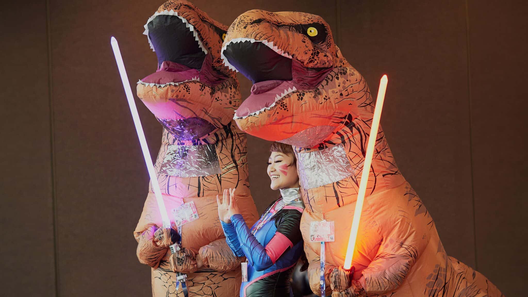 Two individuals in inflatable t-rex costumes wielding lightsabers alongside a smiling person in cosplay return to Sakura-Con, Seattle.