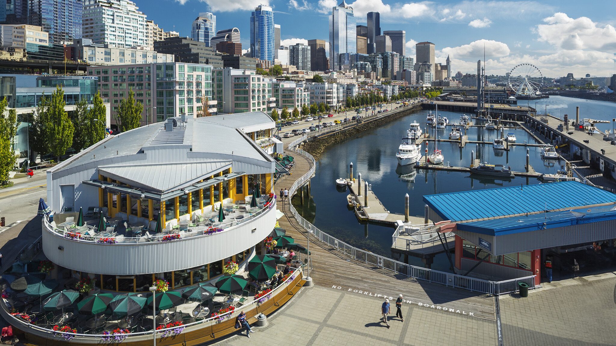 A bustling waterfront with a restaurant terrace overlooking a marina against a backdrop of Seattle's modern city skyline, enhancing tourism.