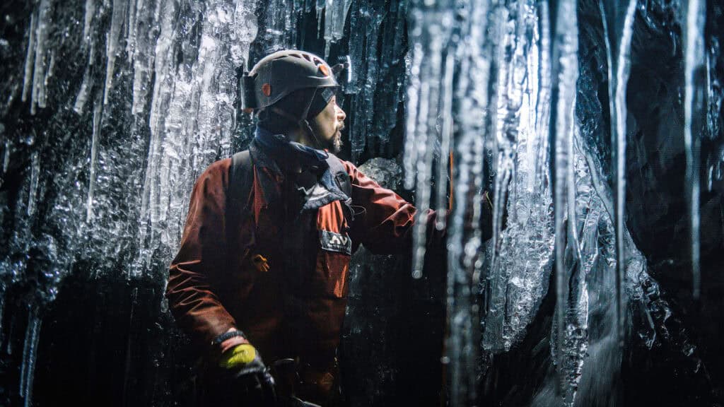 A person wearing a helmet and protective gear explores an icy cave, surrounded by large icicles and ice formations.