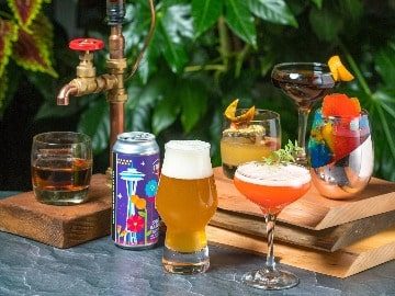 Variety of beverages including Sheringham Distillery cocktails, beer, and a soft drink can, displayed on a wooden table amid lush greenery.