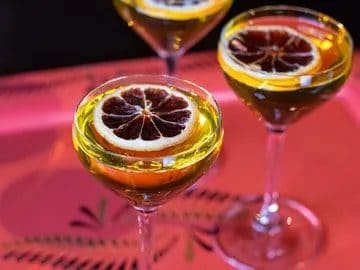 Three cocktail glasses with a slice of dried blood orange on top, set on a pink surface with a patterned design, illuminated by warm lighting at a culinary retreat.