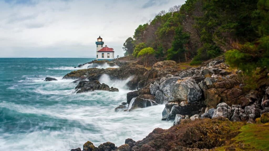 A lighthouse on a rocky coastline with waves crashing against the shore under a cloudy sky, perfect for island hopping destinations.