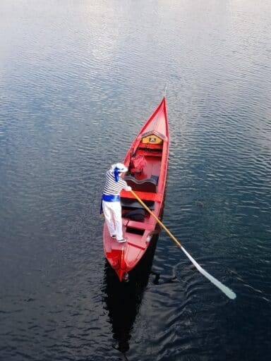 A person in a striped shirt rows a red canoe on a calm blue water surface in California, viewed from above.