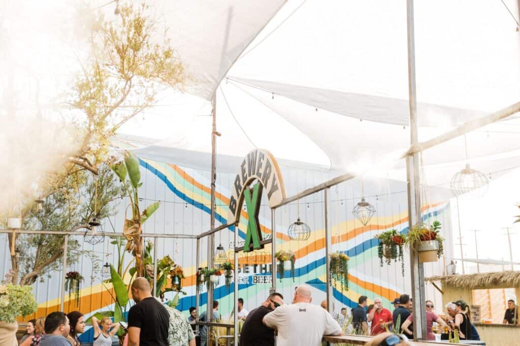 People enjoying drinks at an outdoor brewery in California with colorful, striped walls and hanging plants under a tent.