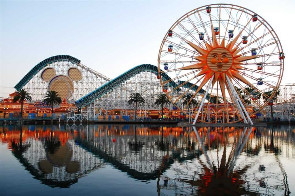 A vibrant amusement park in California, featuring a ferris wheel and roller coaster, reflected in a calm waterfront at sunset.