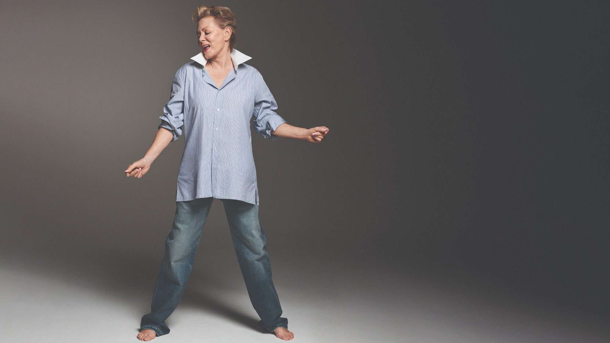 A middle-aged actress dancing joyfully in a studio, wearing jeans and an oversized shirt, with a subtle smile on her face.