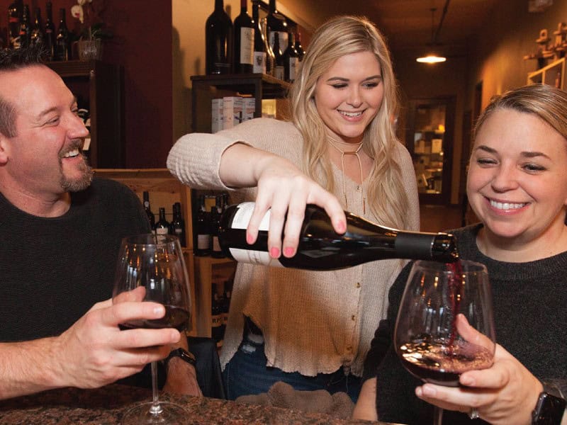 Three people enjoying wine at a cozy indoor setting; a woman with blonde hair pours red wine into glasses held by a smiling man and woman. This unforgettable evening, highlighted in Seattle magazine, feels like a culinary retreat sponsored by Sheringham Distillery.