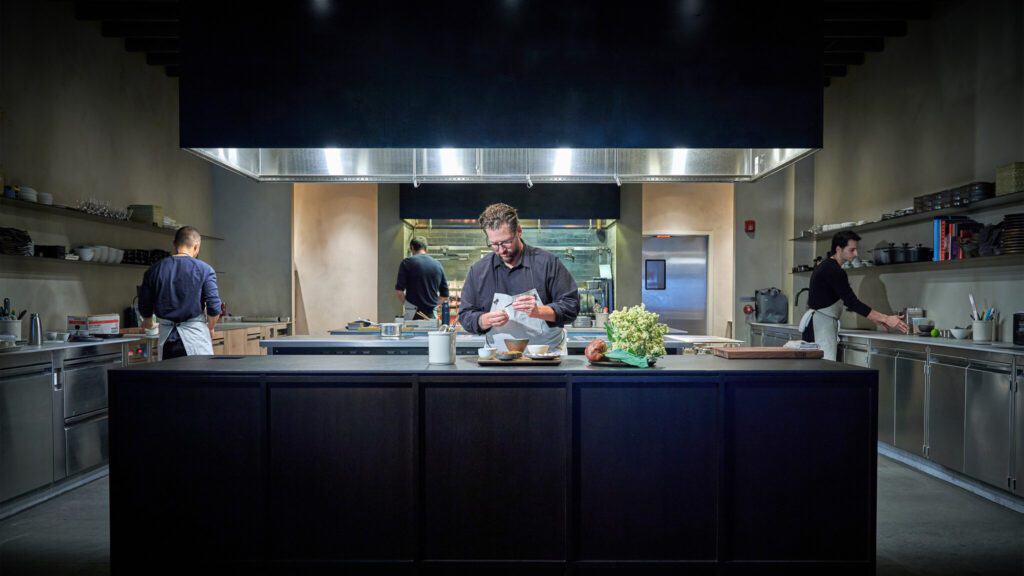 A professional chef focuses on preparing a dish in a modern farmhouse kitchen with two assistants working in the background.