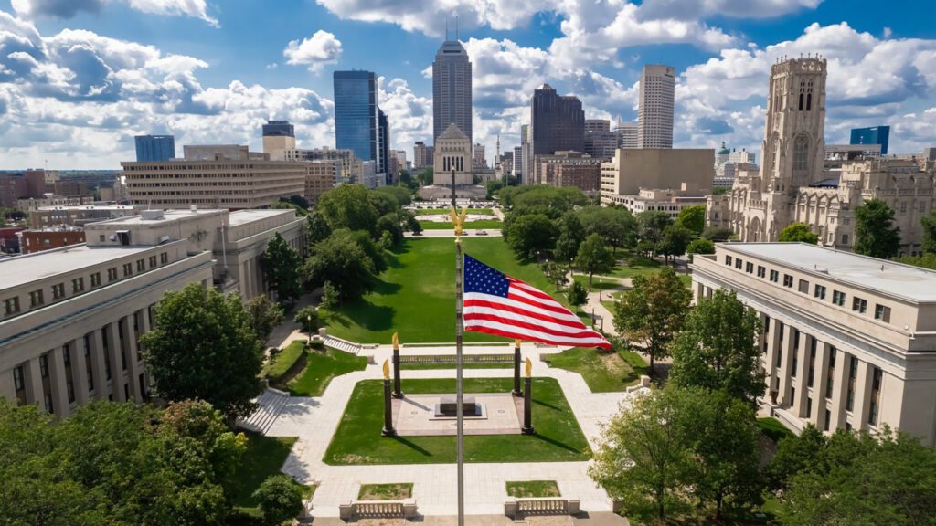 Aerial view of a cityscape with an american flag in the foreground, flanked by classical buildings and a green lawn, with skyscrapers in the background under a partly cloudy sky during a solar