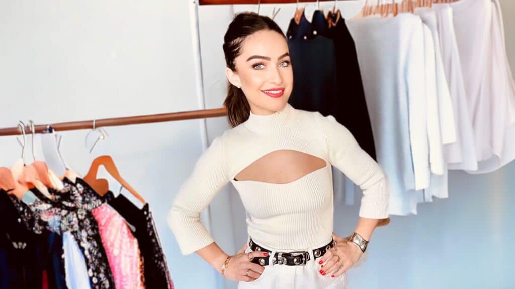 Woman in a stylish white cut-out top and silver belt stands confidently in a room with racks of clothes during her wardrobe update.